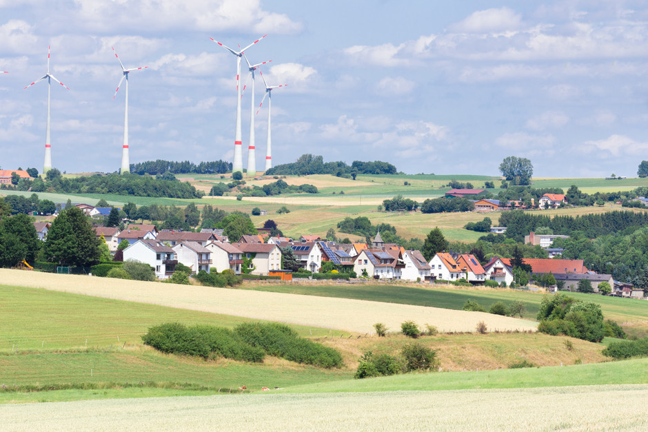 Blick auf ein Dorf in einer ländlichen Gegend. Am Dorfrand stehen fünf Windräder.